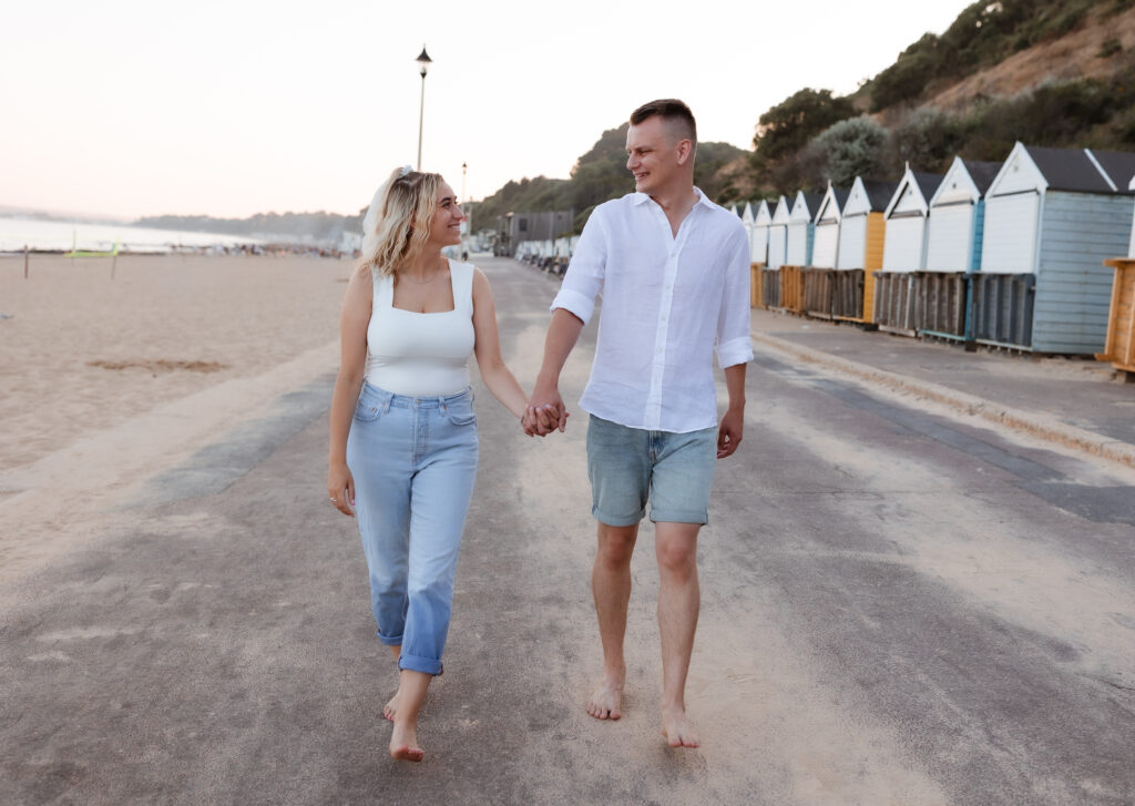 A couple holding hands and smiling while walking barefoot on a beachside path, with colorful beach huts and trees in the background.
