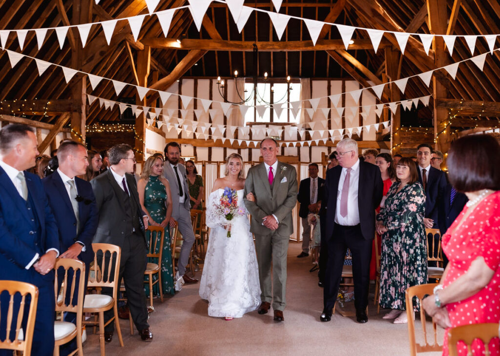 A bride in a white dress walks down the aisle with an older man in a tan suit, surrounded by seated guests in a rustic, decorated barn with white bunting overhead.
