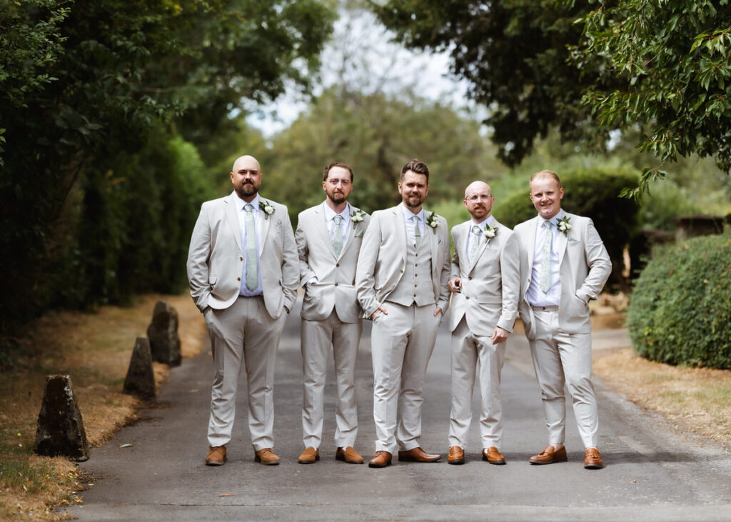 Five men in matching light-colored suits and brown shoes stand together on a tree-lined road, posing for a group photo, possibly at a wedding. They are smiling, and greenery surrounds them on both sides.