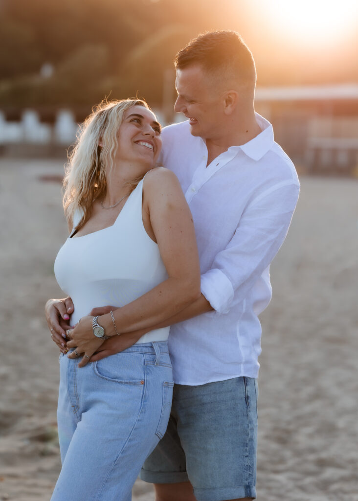 A couple stands on a sandy beach at sunset, smiling and embracing each other warmly, with the golden sun glowing in the background.