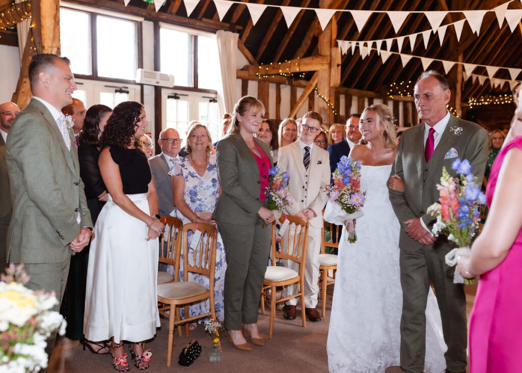 A bride in a white dress walks down the aisle with an older man. Guests smile and watch. The groom waits at the front. Colorful flowers and bunting decorate the bright wedding venue.