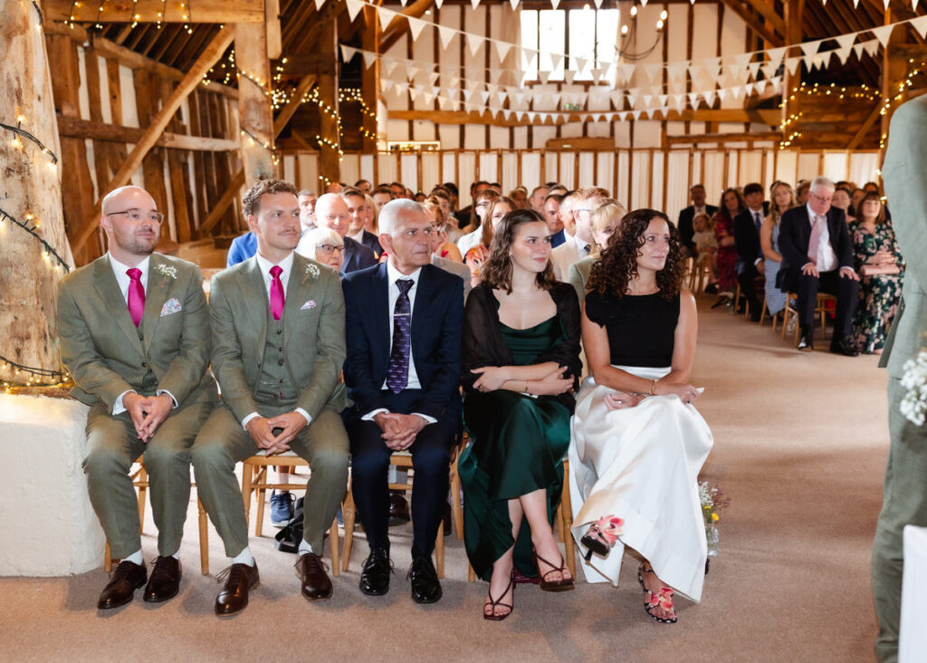 A group of people, dressed formally, sit in the front row at a rustic indoor wedding ceremony, with fairy lights and bunting above, and more guests seated in the background.
