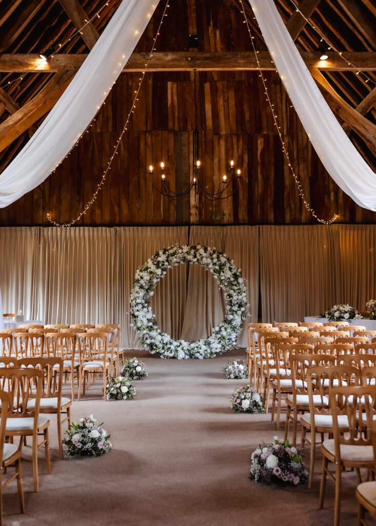 A rustic wedding venue with wooden beams, draped white fabric, hanging string lights, wooden chairs, floral arrangements lining the aisle, and a large circular floral arch at the front.
