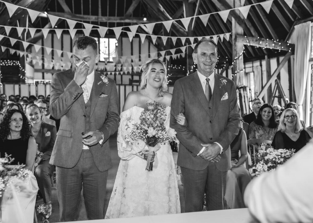 A bride in a white dress stands smiling between two men in suits; one wipes his eyes. They are in a decorated hall with guests seated behind them, suggesting a wedding ceremony.