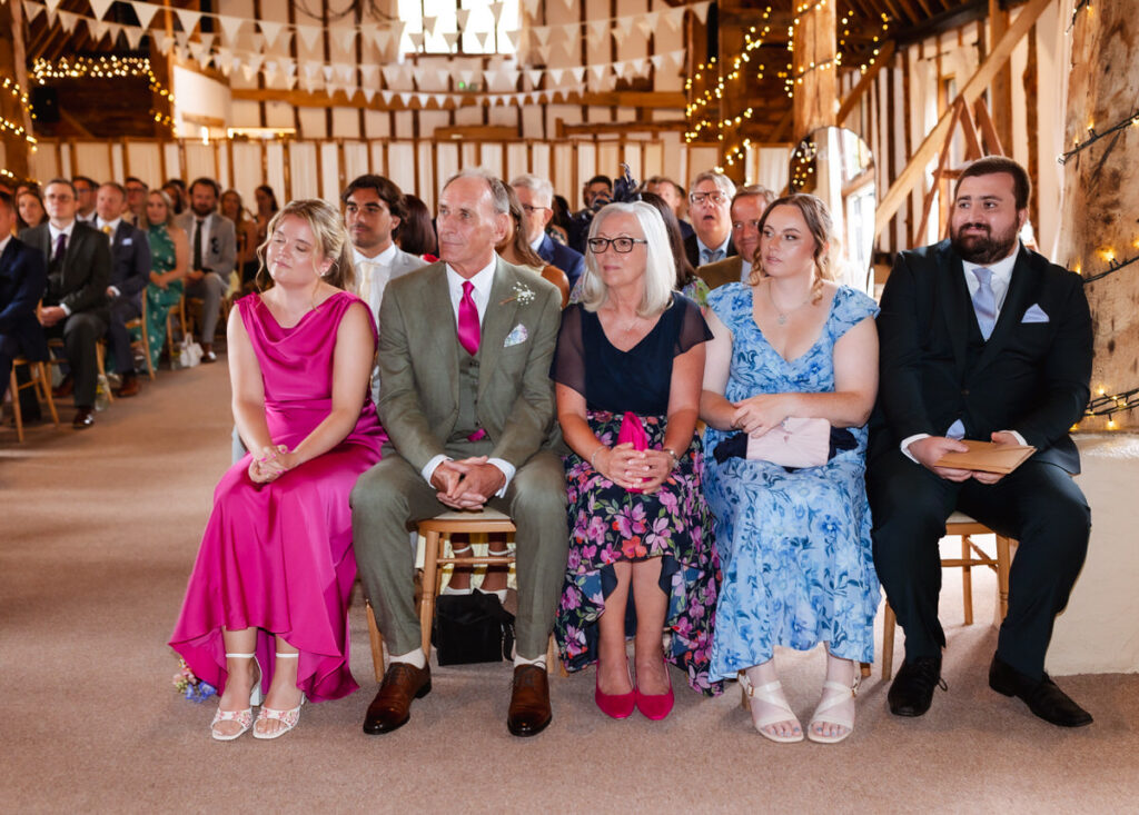 A group of well-dressed people sit in rows at a decorated indoor wedding venue, with fairy lights and bunting overhead. The front row features four adults and one young woman in colorful outfits, looking toward the ceremony.
