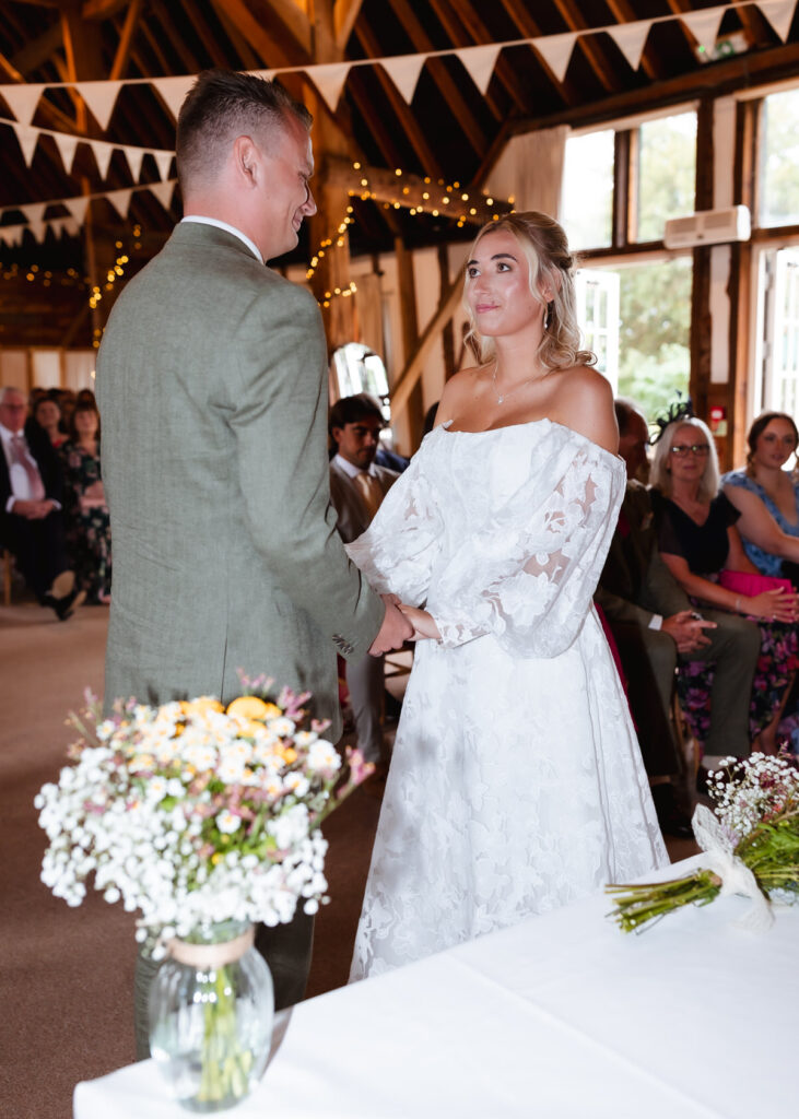 A bride in a white lace dress and a groom in a green suit stand holding hands during their wedding ceremony in a rustic, decorated hall, with guests seated behind them.