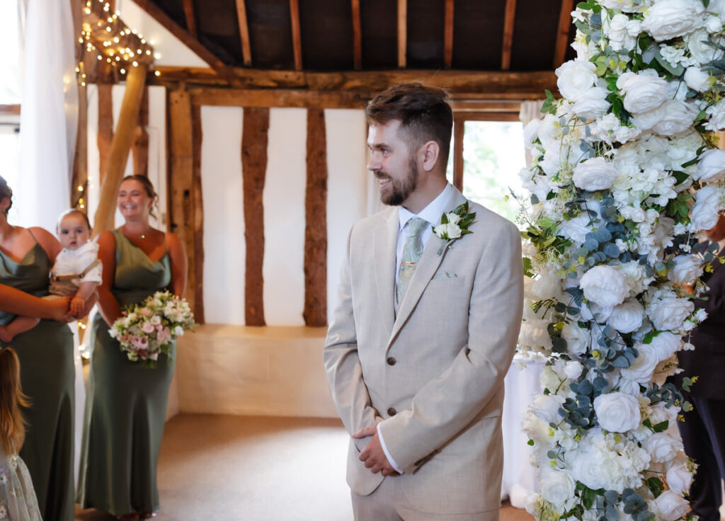 A groom in a beige suit smiles as he stands by a floral arch. Bridesmaids in green dresses, one holding a baby, are in the background. The setting is indoors with rustic wooden beams and white flowers.