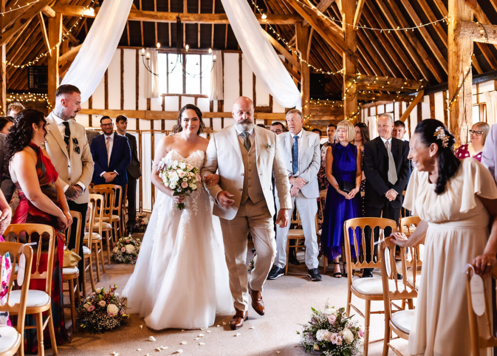 A bride in a white dress walks down the aisle with an older man in a suit, surrounded by guests seated in a rustic barn decorated with flowers and fairy lights.