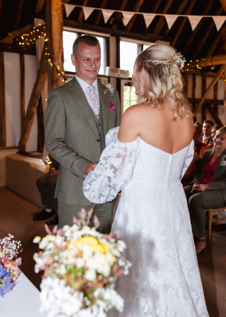 A bride and groom stand facing each other, holding hands during their wedding ceremony in a rustic, decorated venue with guests seated in the background.
