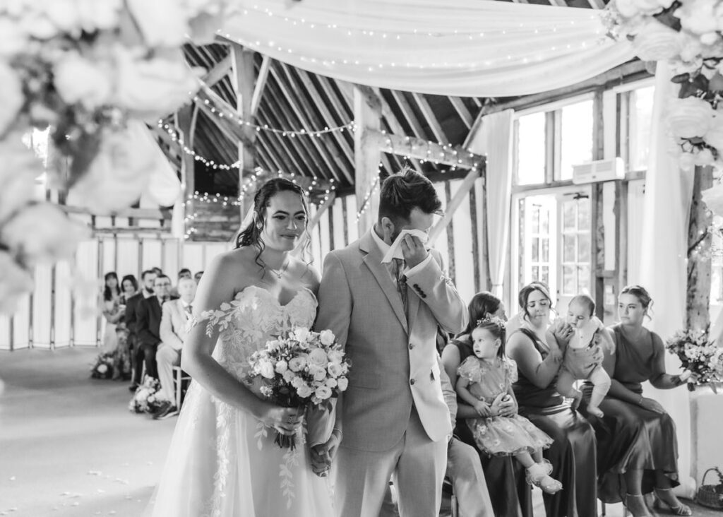 A bride and groom walk down the aisle; the groom wipes away tears while holding the brides hand. Guests seated in the background watch. The venue is decorated with flowers and draped lights.
