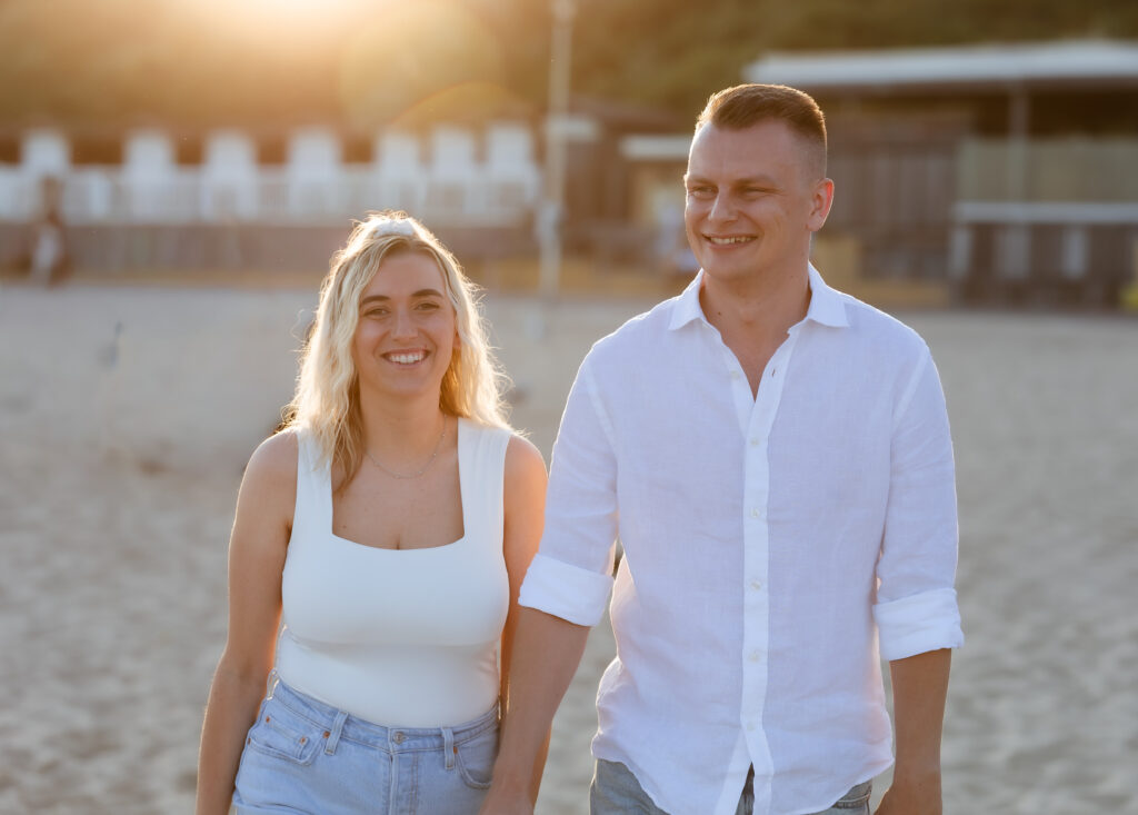 A smiling couple walks hand in hand on a sandy beach at sunset, both wearing casual white tops. The sun shines behind them, creating a warm, golden glow.