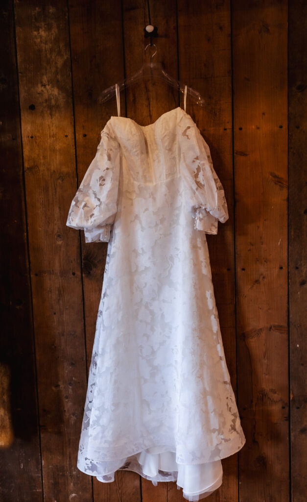 A white lace wedding dress with puffed sleeves hangs on a clear hanger against a rustic wooden wall.
