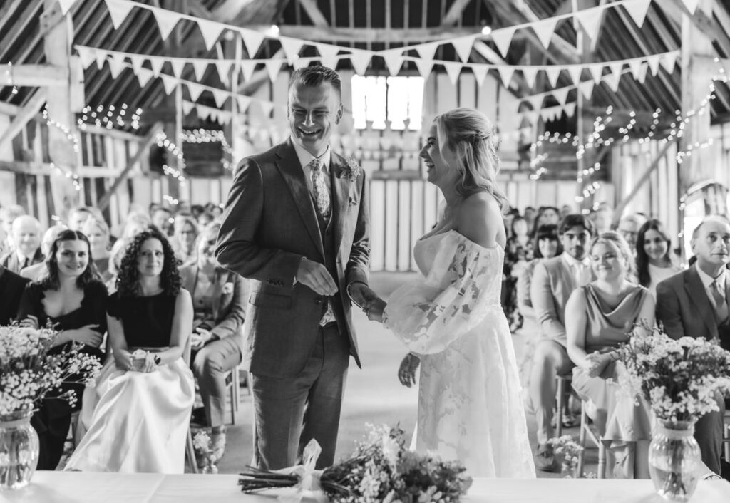 A bride and groom smile at each other during their wedding ceremony in a rustic, decorated barn, surrounded by seated guests, with flowers and bunting visible.