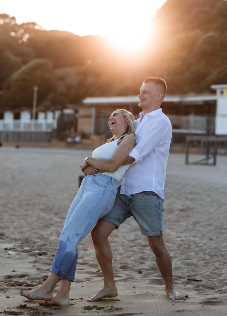 A couple stands barefoot on a sandy beach at sunset, laughing joyfully as the man hugs the woman from behind. The sunlight glows warmly, and trees and buildings are visible in the background.