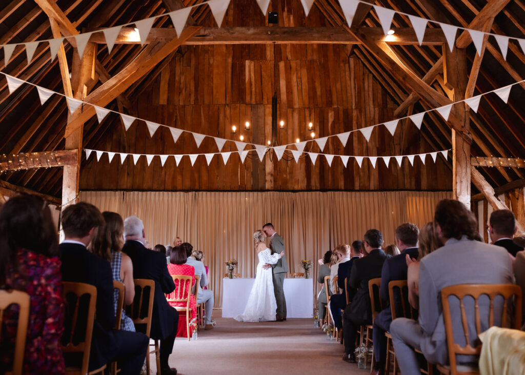 A bride and groom share a kiss at the altar in a rustic barn venue, surrounded by seated guests. White bunting and wooden beams decorate the space, creating a warm, festive atmosphere.