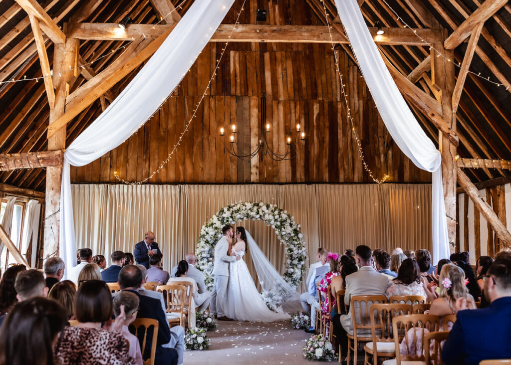 A bride and groom kiss under a floral arch in a rustic barn, surrounded by seated guests. White drapes and string lights decorate the space, creating a romantic wedding atmosphere.
