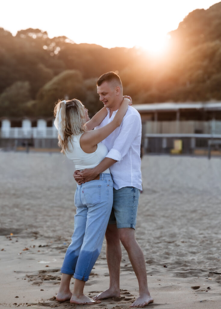 A couple stands barefoot on a sandy beach at sunset, embracing and smiling at each other with trees and buildings in the background.