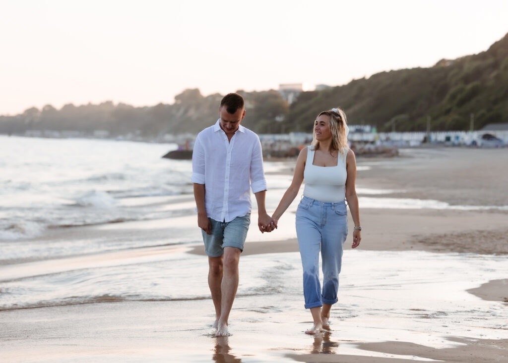 A man and woman walk hand in hand along a beach at sunset, smiling and talking. The shoreline and hills are in the background, with gentle waves by their feet.