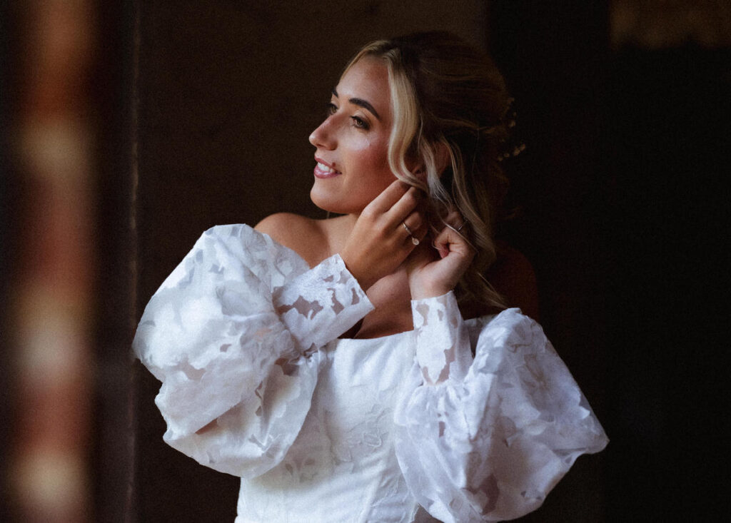 A woman in a white, off-the-shoulder dress with puffy sleeves adjusts an earring, looking to the side and smiling softly in a softly lit setting.