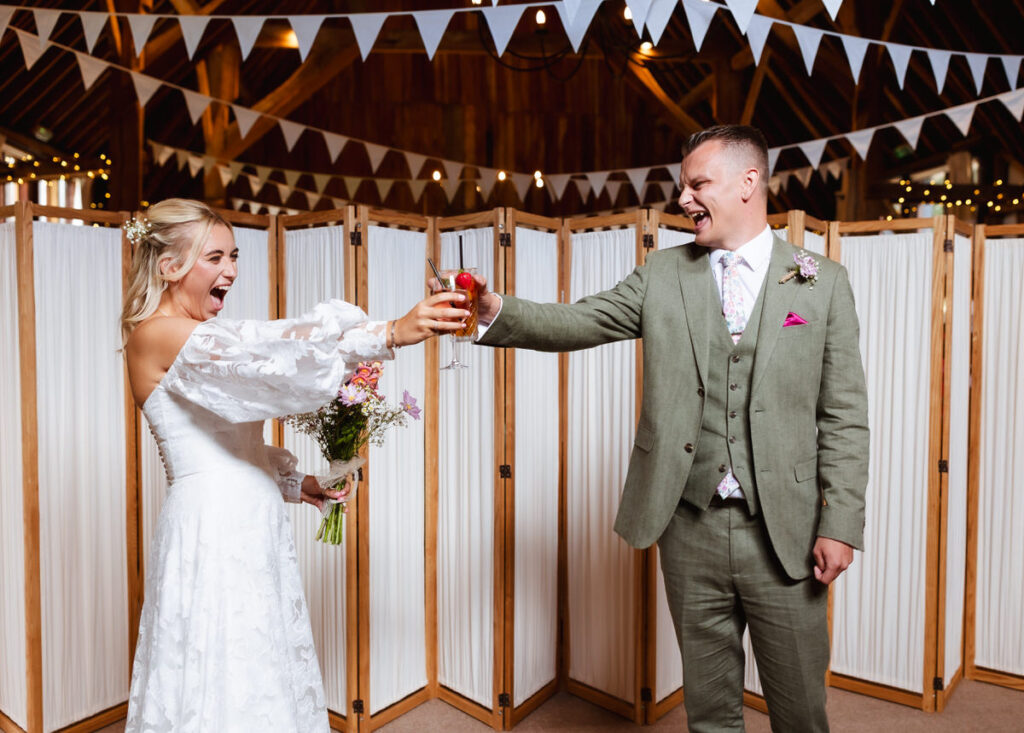 A bride and groom cheerfully clink glasses at their wedding, smiling widely. The bride holds a bouquet, and white bunting decorates the festive indoor setting behind them.