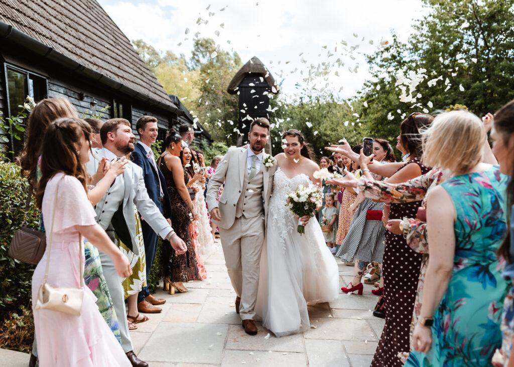 A bride and groom walk down an outdoor path, smiling, as guests throw flower petals in celebration. The couple is surrounded by well-dressed friends and family on a sunny day.