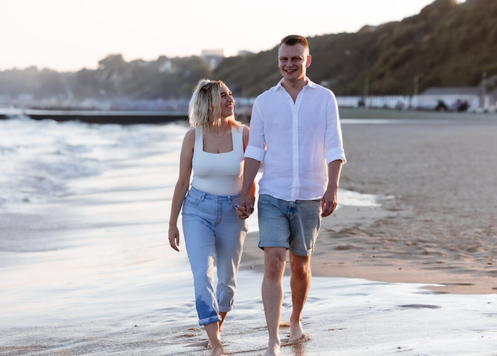 A couple walks barefoot on a sandy beach, holding hands and smiling at each other, with gentle waves and greenery in the background.