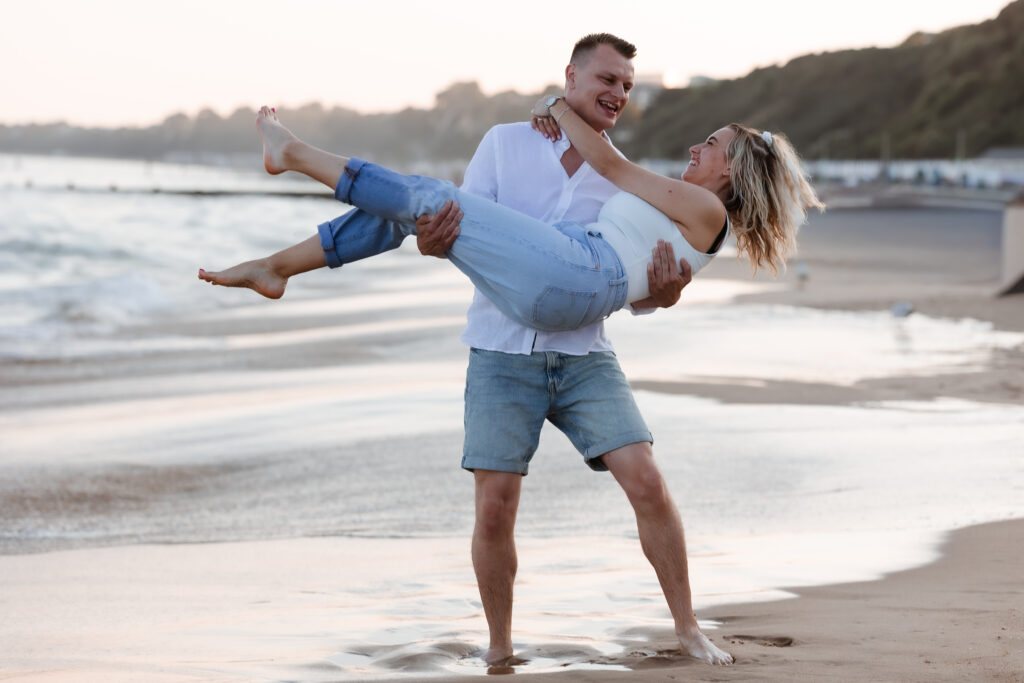 A man in shorts and a white shirt lifts a smiling woman in jeans and a white top on a sandy beach with waves and cliffs in the background. Both appear happy and playful.