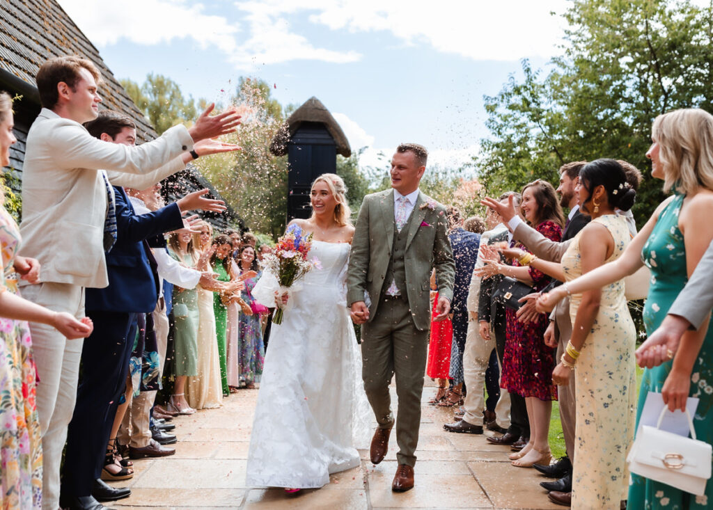 A newlywed couple walks hand in hand down an outdoor aisle as guests on both sides throw confetti and smile. The bride wears a white gown and holds flowers; the groom wears a light suit.