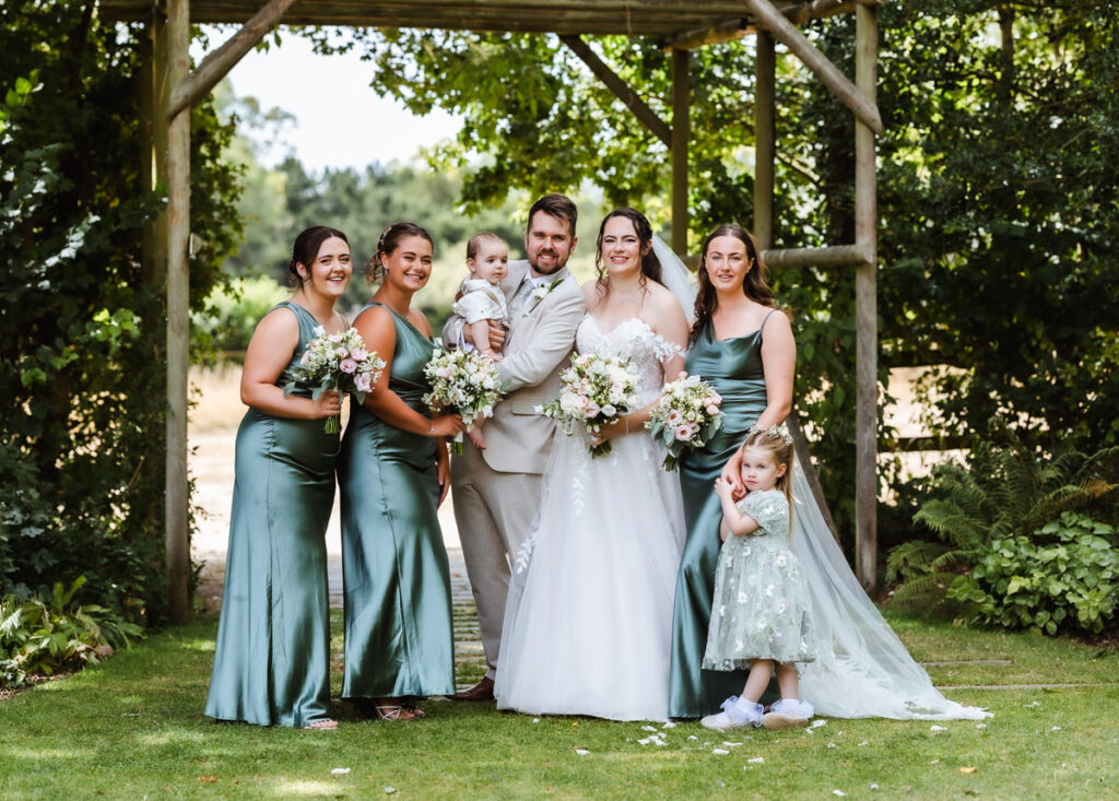 A bride, groom, two young children, and three bridesmaids in green dresses pose smiling together outdoors under a wooden arch surrounded by greenery.