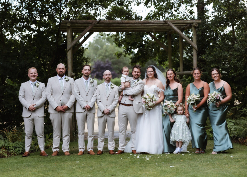 A wedding party poses outdoors under a wooden arch. The bride and groom stand center, the bride holding a bouquet. Bridesmaids wear teal dresses, groomsmen wear light suits. Two children, one flower girl and one baby, are with them.