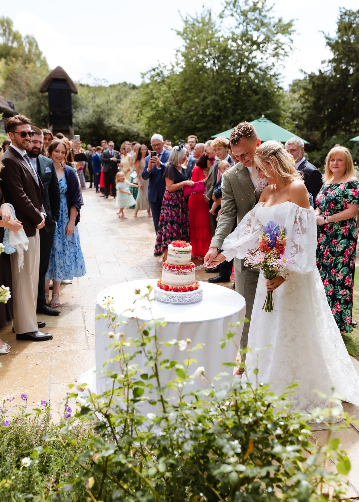A bride and groom cut their wedding cake outdoors, surrounded by guests. The bride holds a bouquet, and everyone is smiling. The scene is bright and festive with greenery and flowers.