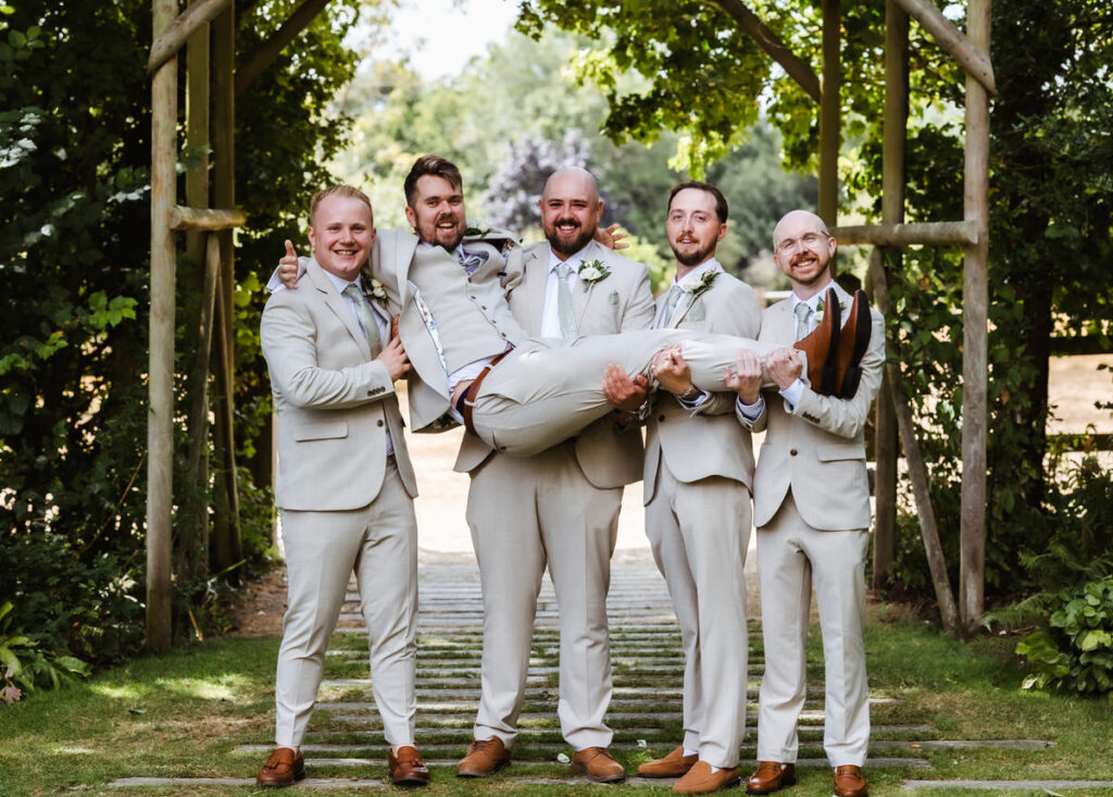Five men in matching light suits pose outdoors; four stand and smile while holding up the fifth man horizontally under a wooden archway, surrounded by greenery.