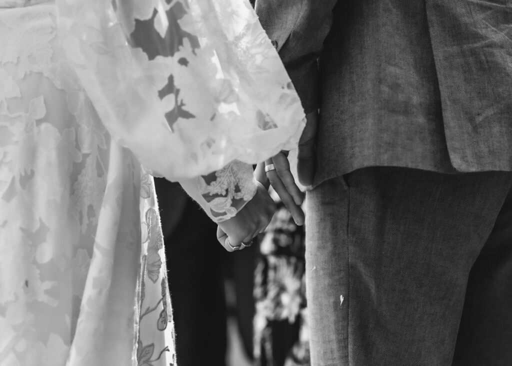 Black and white photo of two people holding hands, one wearing a floral lace dress and the other in a suit, seen from behind, suggesting a romantic or wedding moment.