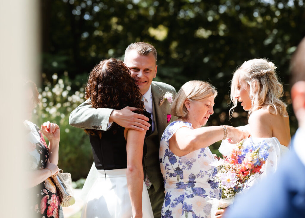 A man in a suit hugs a woman with curly hair, while two other women, one holding a colorful bouquet, stand nearby outdoors in sunlight.