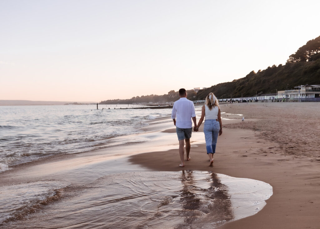 A couple walks hand in hand along a sandy beach at sunset, with gentle waves beside them and hills in the background.