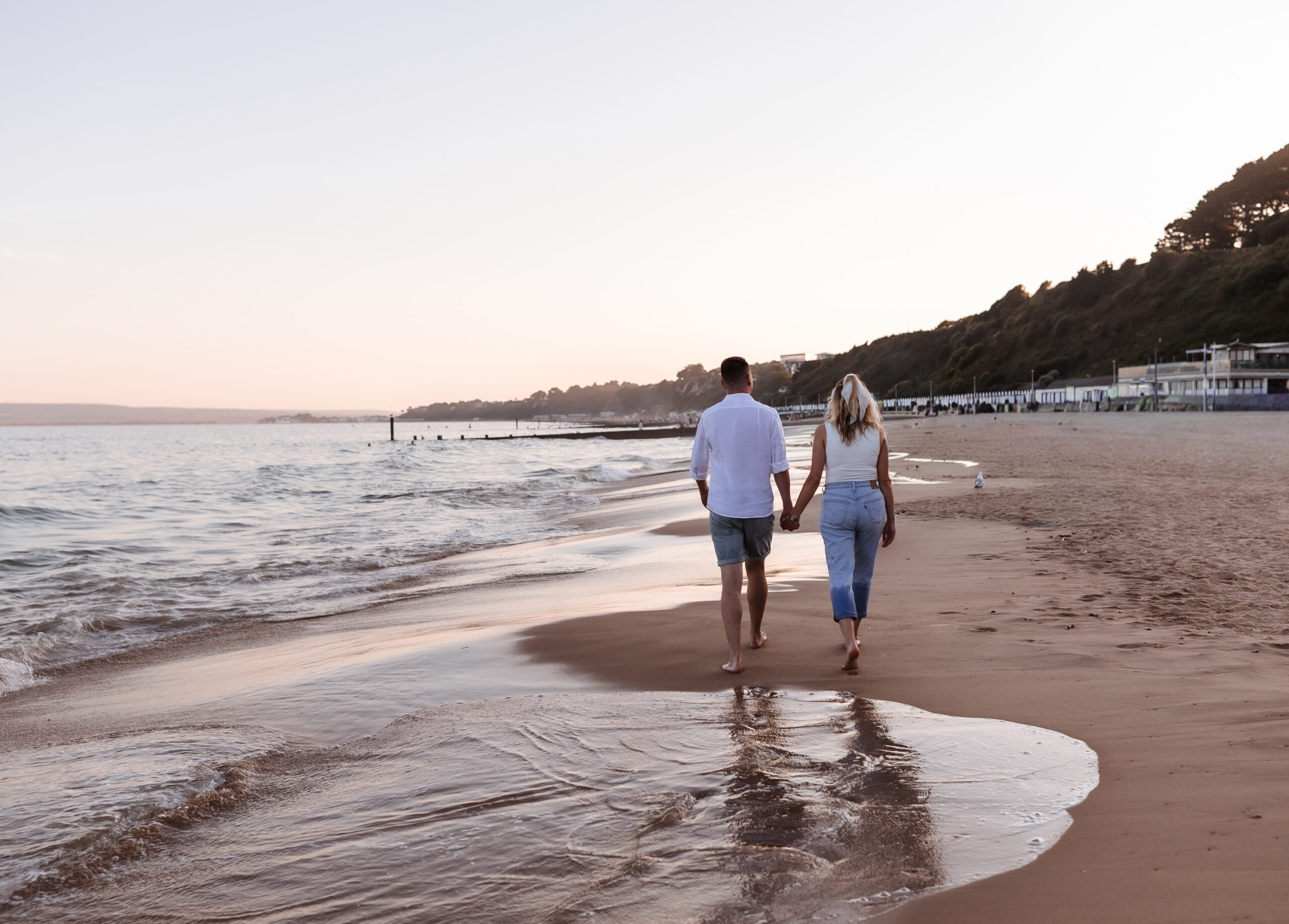 A couple walks hand in hand along a sandy beach at sunset, with gentle waves beside them and hills in the background.