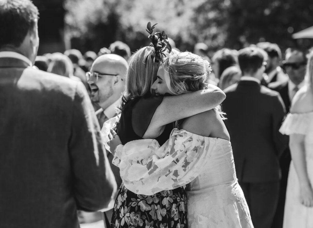 A bride in a white dress hugs a woman in a floral dress at an outdoor wedding, surrounded by smiling and talking guests.