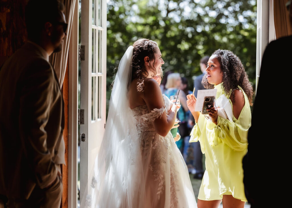 A bride in a white dress talks and laughs with a guest in a yellow dress by an open door, sunlight streaming in and people visible outside in the background.