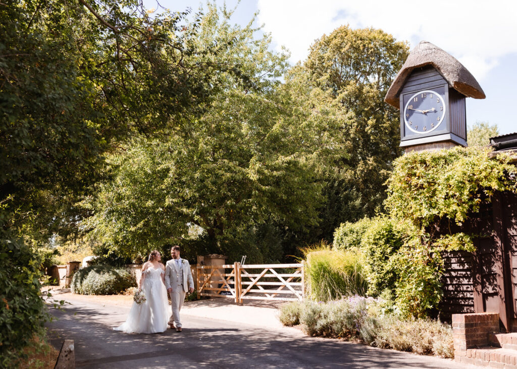 A bride and groom walk hand in hand down a sunlit lane beside a rustic building with a thatched clock tower, surrounded by lush greenery and trees.