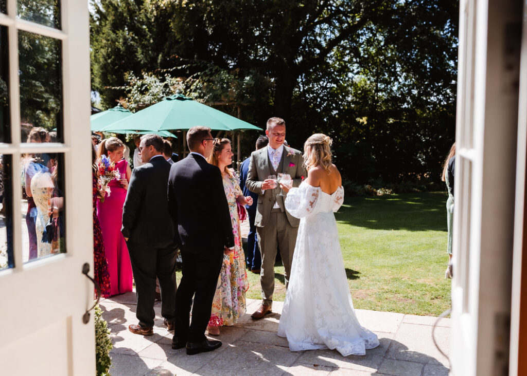 A bride in a white dress talks with guests in formal attire outside under sunlight, with a green umbrella and trees in the background, seen from an open doorway.