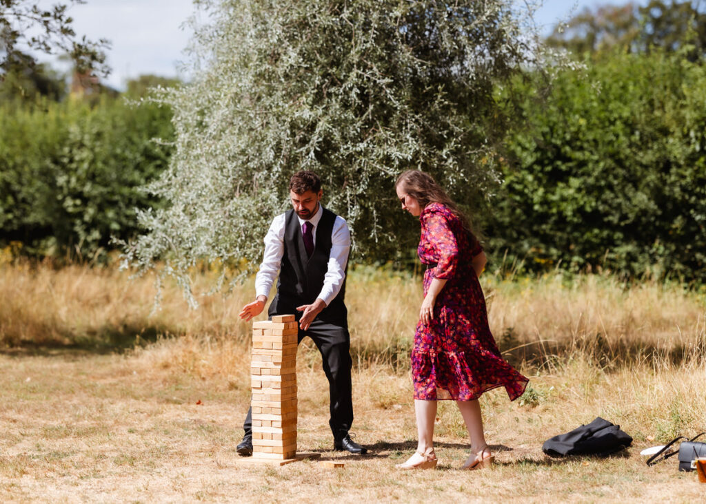 A man in a suit and a woman in a red floral dress play a giant Jenga game outdoors on a sunny day, surrounded by trees and tall grass.