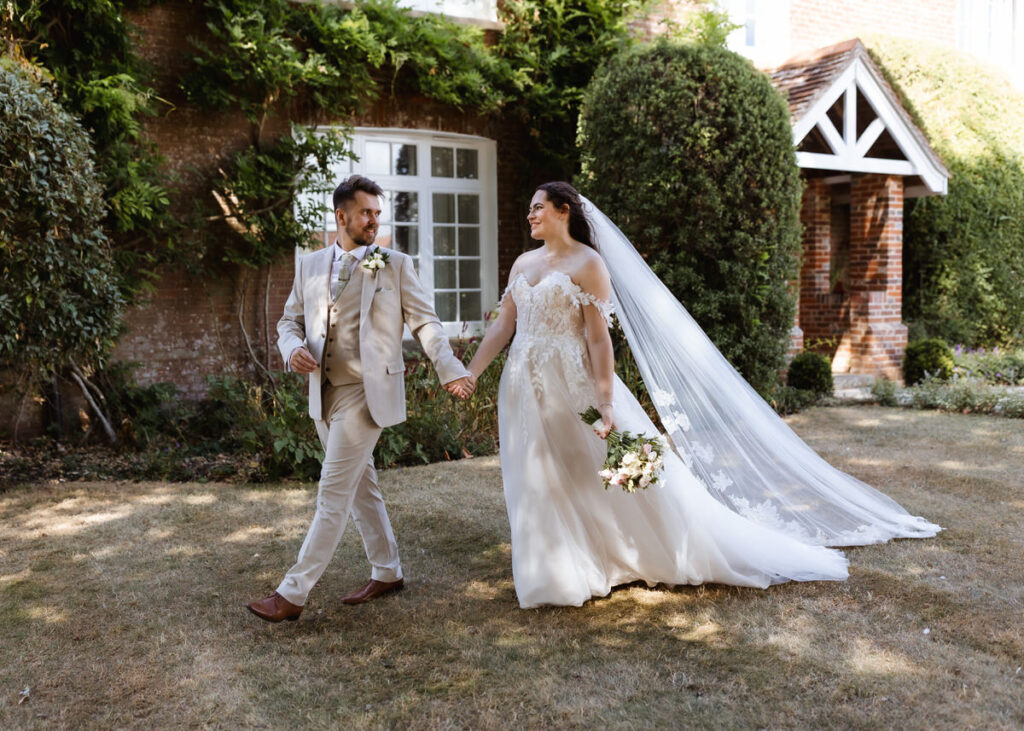 A bride and groom walk hand in hand on grass outside a brick house. The bride wears a white dress with a long veil, holding a bouquet. The groom wears a beige suit and boutonniere. Both are smiling.