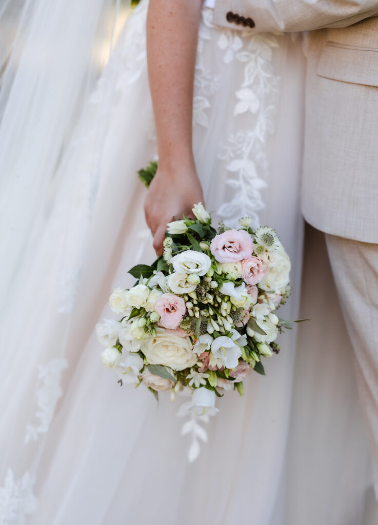 A bride in a white floral lace dress holds a bouquet of white and pale pink flowers, standing beside someone in a light beige suit.