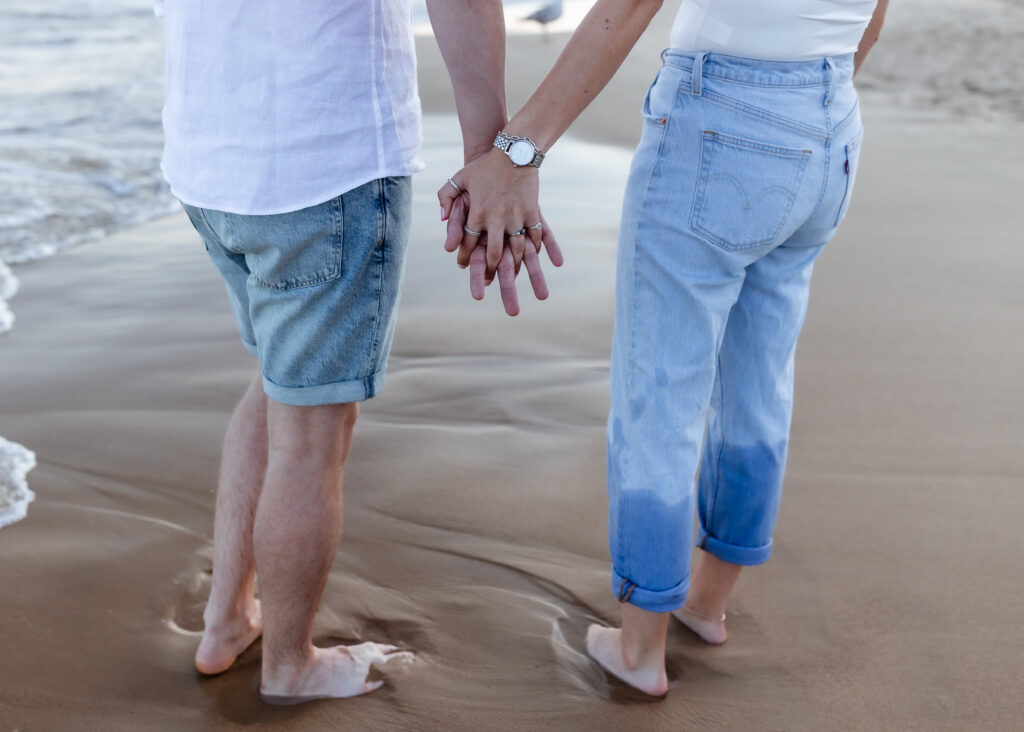 A couple holding hands while standing barefoot on wet sand at the beach, with gentle waves nearby.