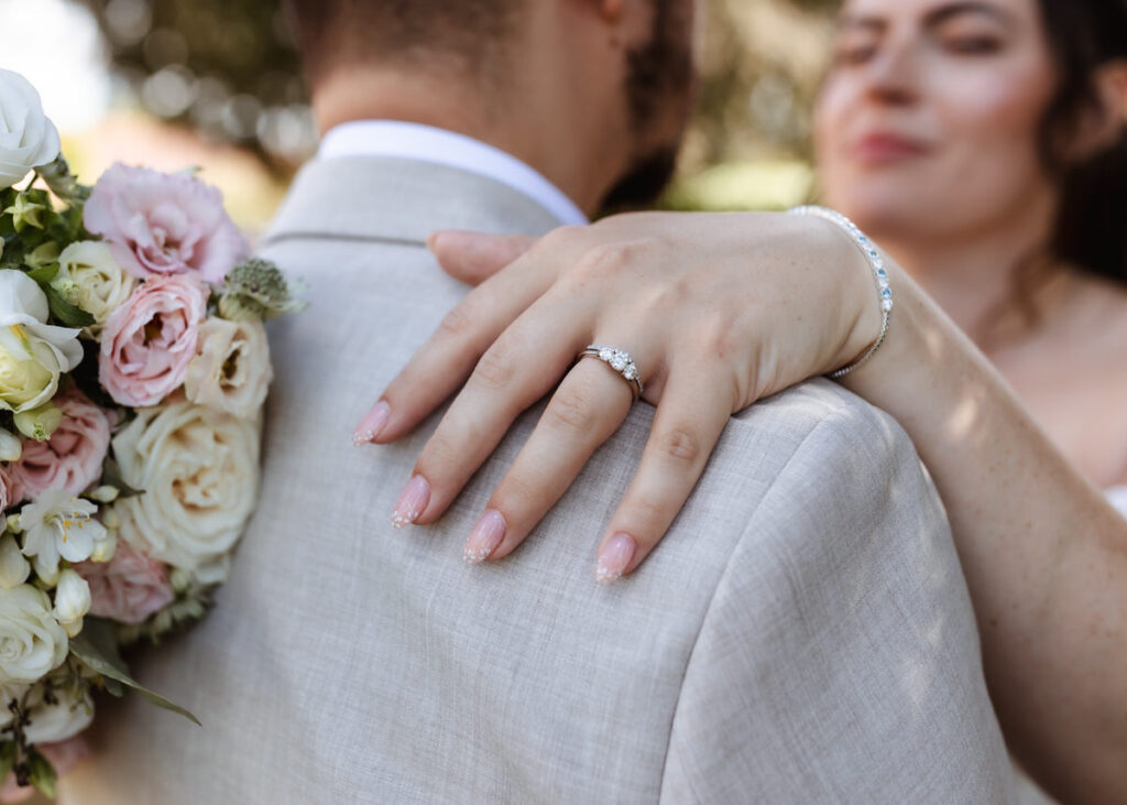 A womans manicured hand with a diamond ring rests on a mans shoulder. She holds a bouquet of pale pink and white flowers. Both wear light-colored attire, and their faces are softly out of focus.