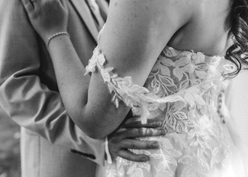 A close-up black and white photo of a bride and groom embracing, showing the brides lacy dress and the grooms suit, with their arms gently wrapped around each other.