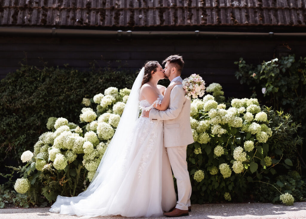 A bride and groom kiss in front of blooming white hydrangeas, with the bride in a white gown and veil and the groom in a light-colored suit, outdoors on a sunny day.