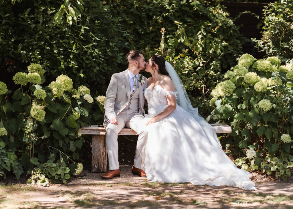 A bride and groom share a kiss while sitting on a wooden bench in a garden, surrounded by lush green hydrangeas and foliage. Both are dressed in wedding attire and look happy.