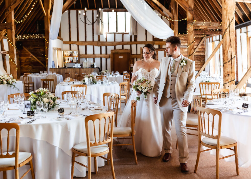 A bride and groom walk hand in hand through a rustic wedding venue with round tables, white tablecloths, floral centerpieces, and wooden beams decorated with string lights.