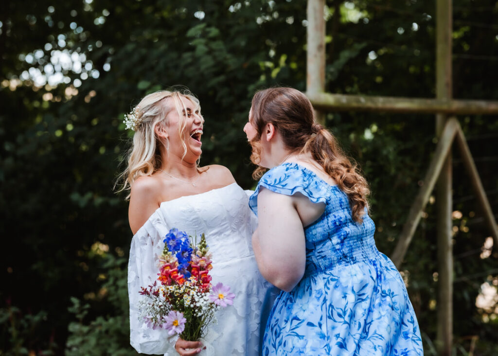 Two women outdoors laughing together; one wears a white dress holding a colorful bouquet, the other wears a blue floral dress. They face each other joyfully with greenery in the background.
